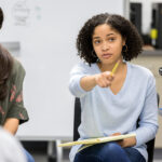 A serious female high school student sits in a circle with a group of classmates and takes a questions as she leads a study group discussion.  She uses her pencil to point to an unrecognizable classmate.